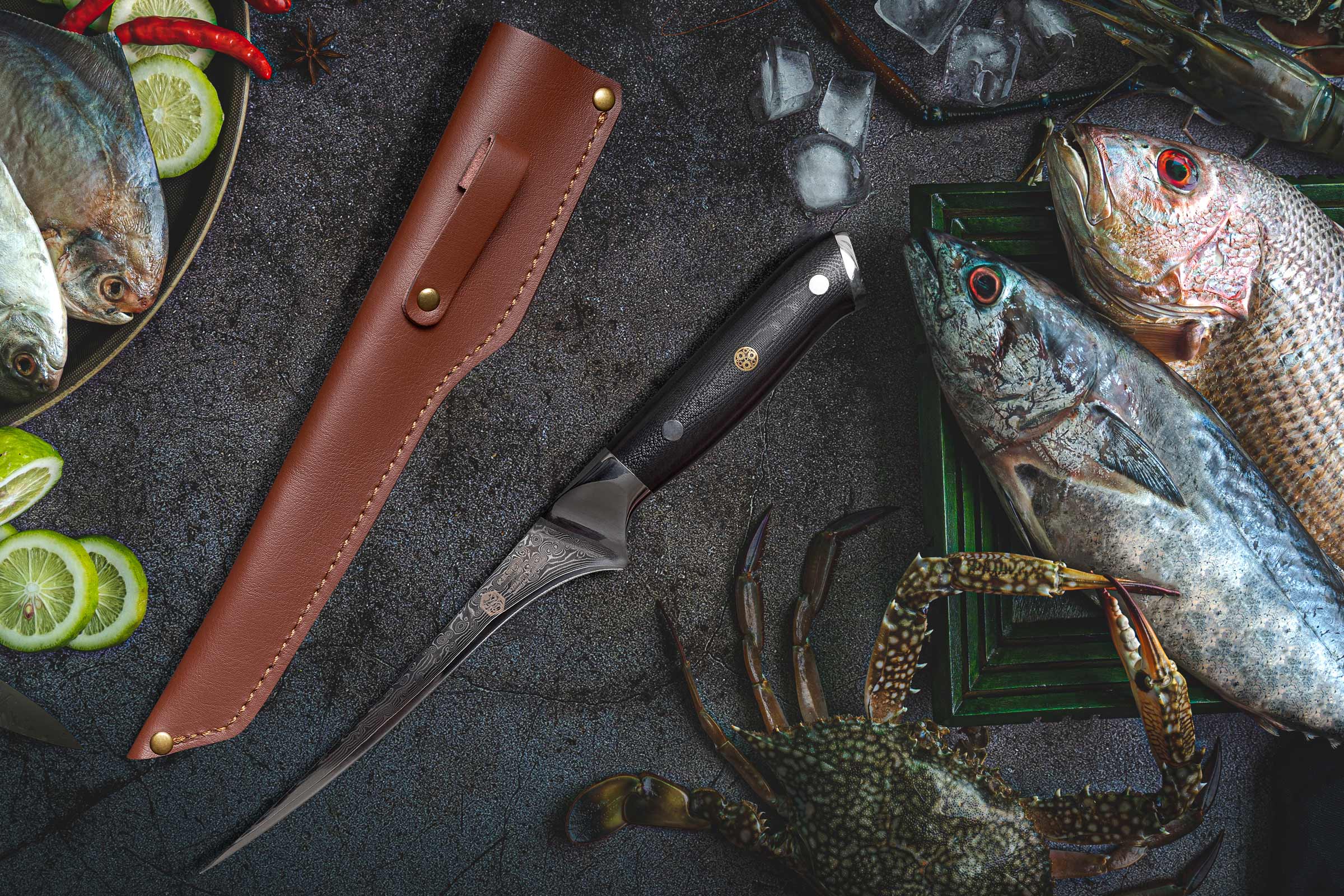 The Kessaku Leather Fillet Knife Sheath with the Dynasty Damascus Fillet Knife surrounded by fish, crab, and slices of lime.