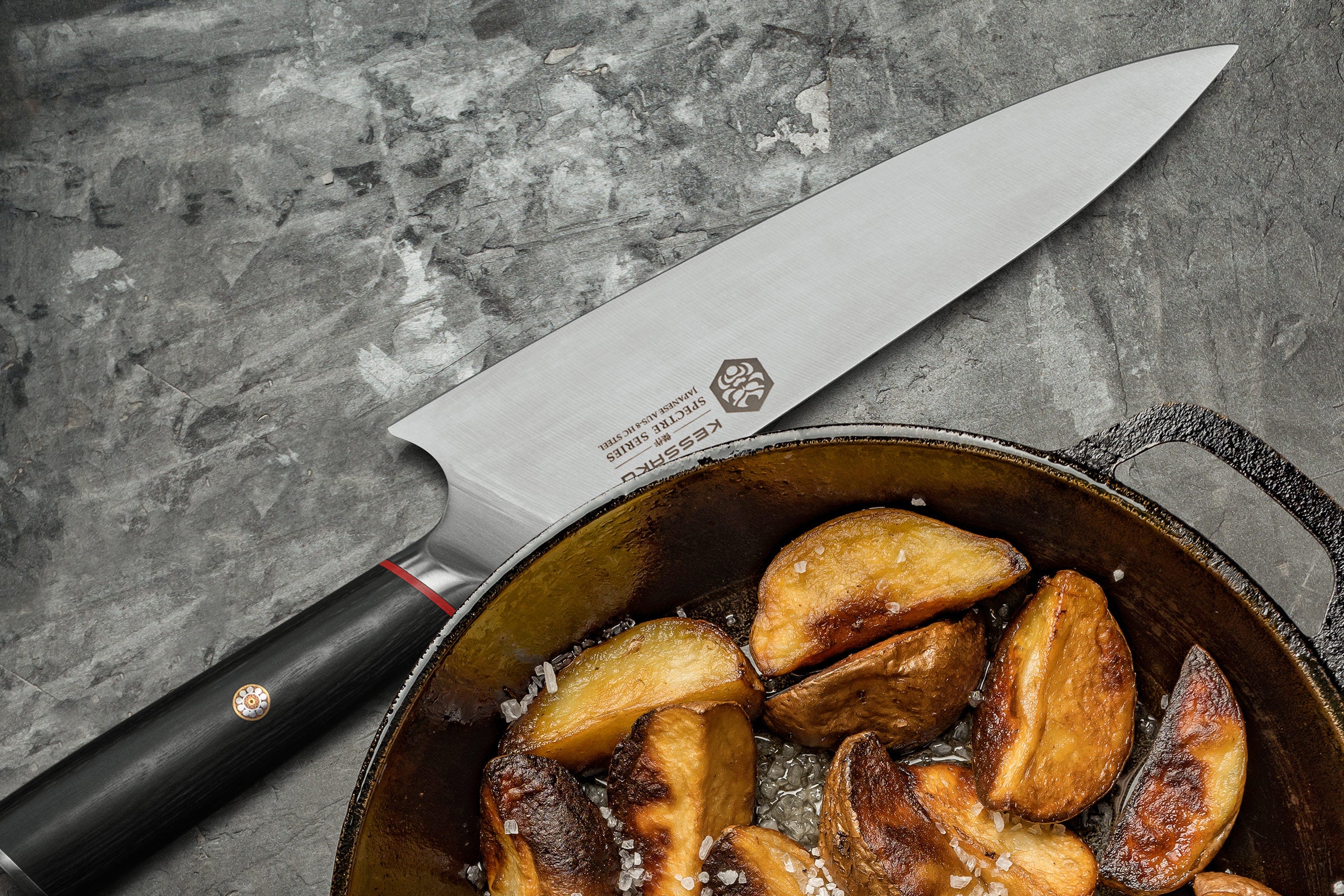 The Spectre Chef's Knife next to a skillet of potatoes wedges frying.