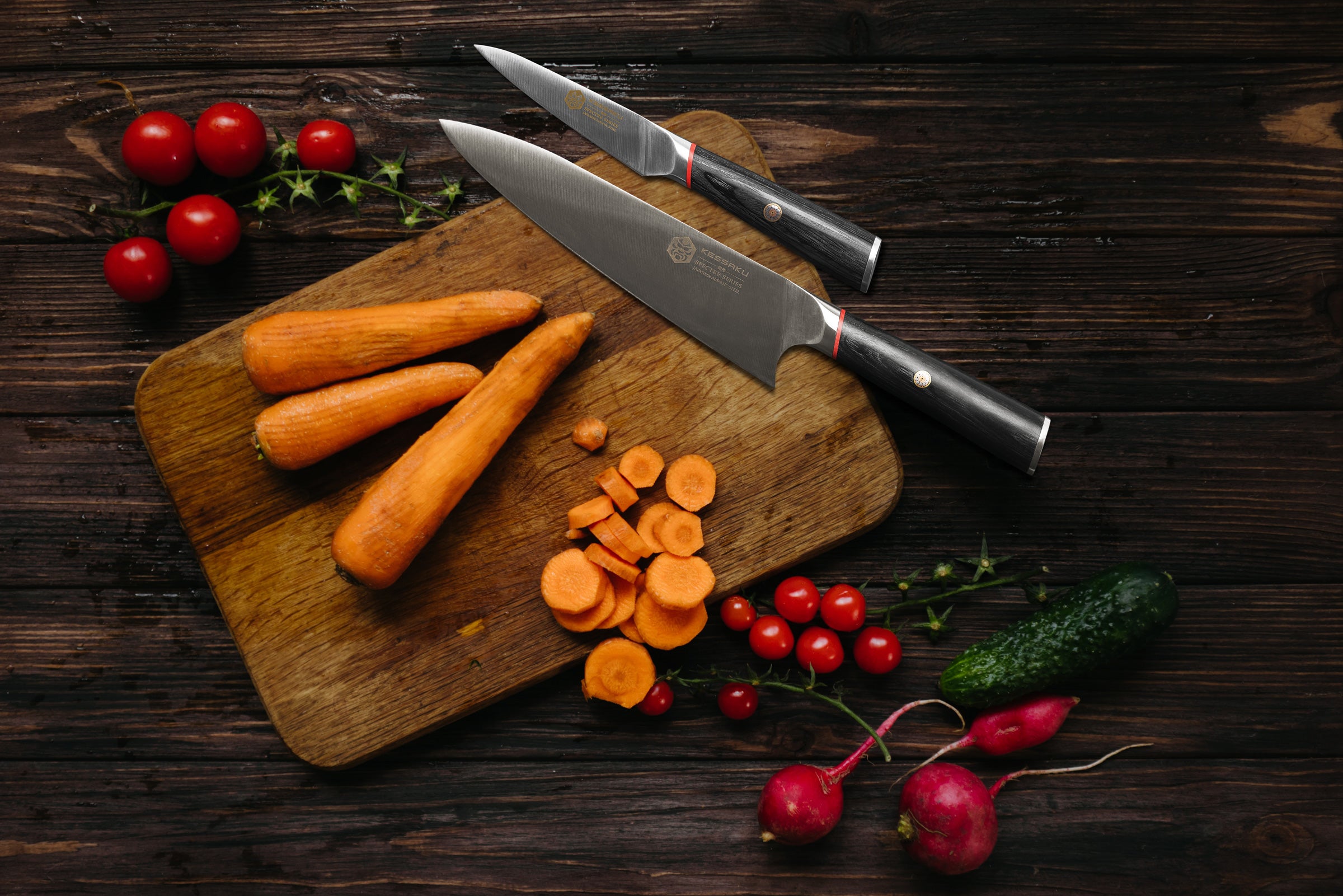 The Spectre Chef's Knife and Paring Knife on a cutting board with peeled and sliced carrots, surround by radishes, and a pickle.