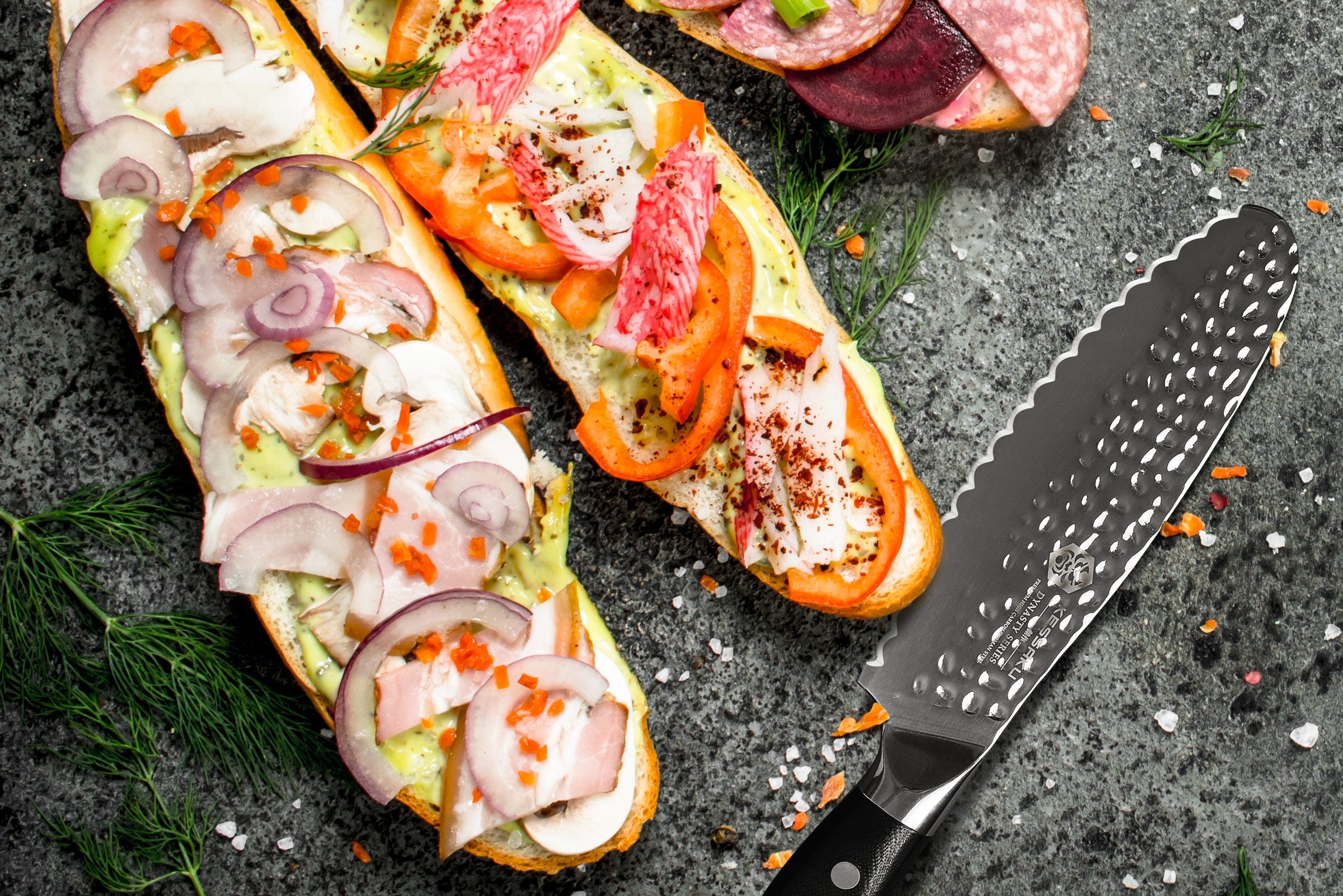 The Dynasty Serrated Sandwich Knife having just prepped three sandwiches with ingredients such as red peppers, red onions, and cured meats.