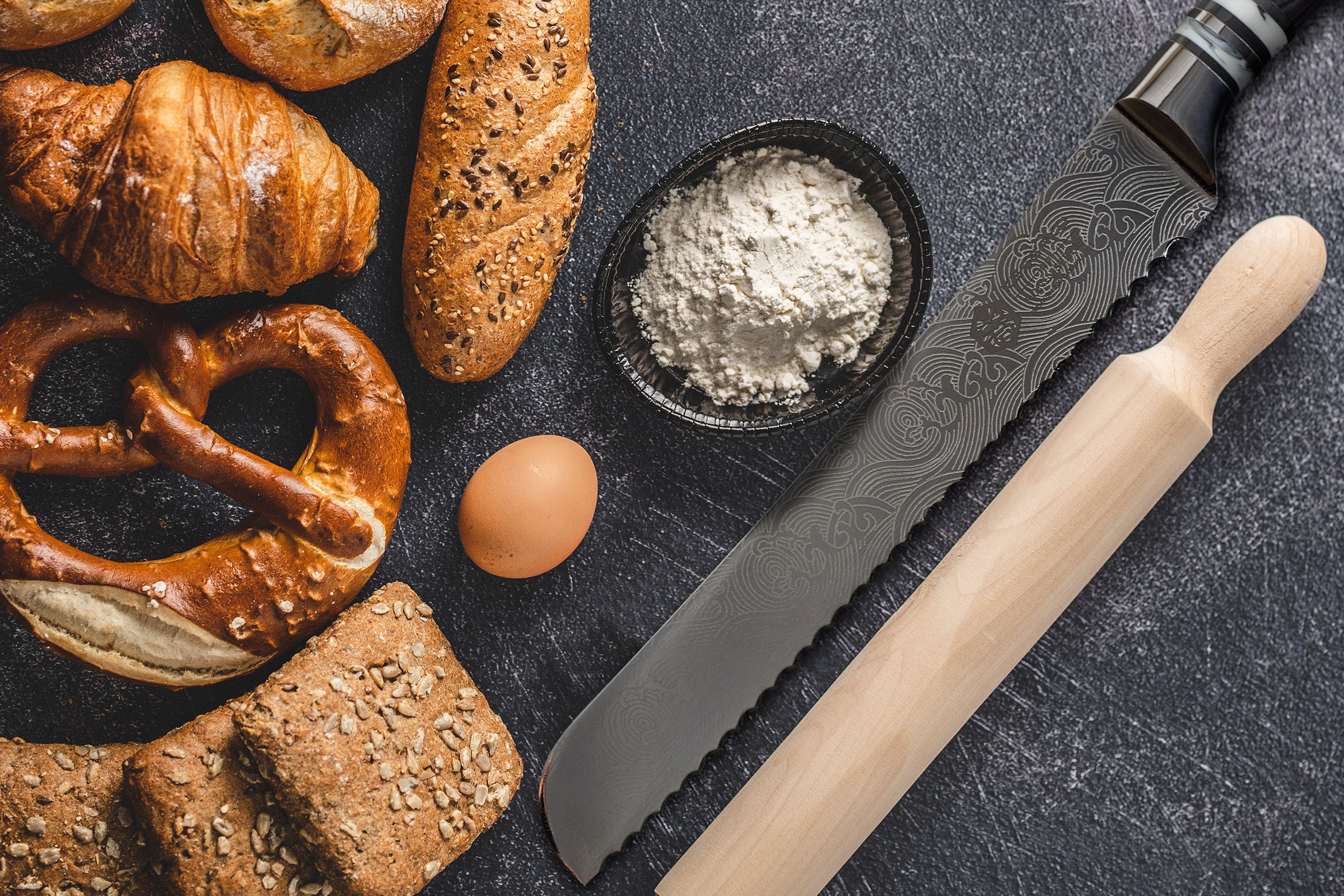 The Ronin Bread Knife next to a rolling pin, cup of flour, egg, whole grain breads, homemade pretzel, and croissants. 