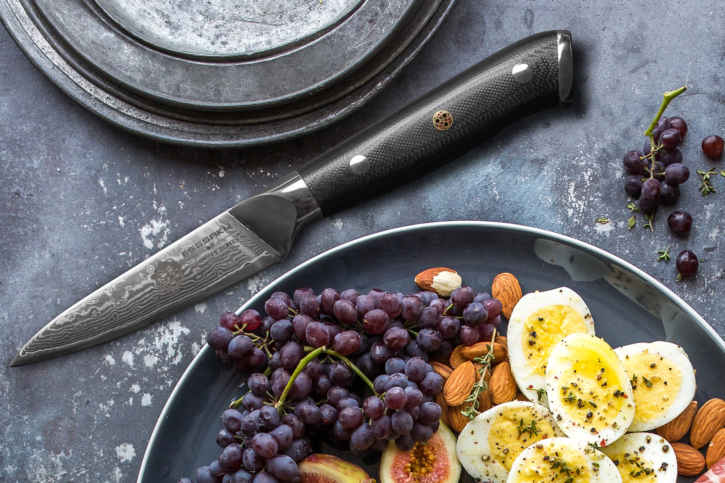 The Dynasty Damascus Paring Knife with a plate of sliced hard boiled eggs, figs, red grapes, and almonds.
