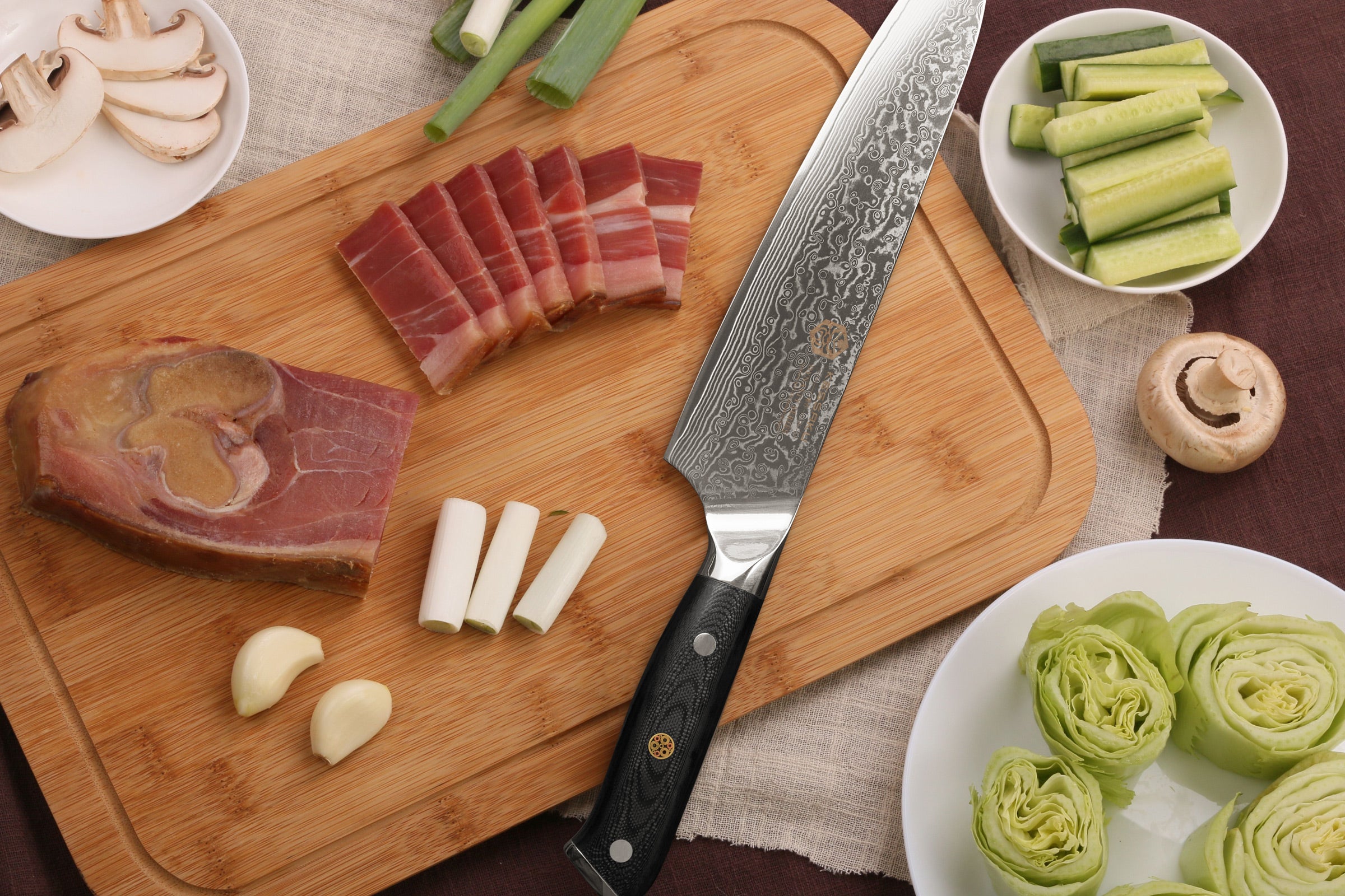 The Kessaku Dynasty Damascus Chef's Knife with thick slices of bacon, green onion, cloves of garlic, zucchini